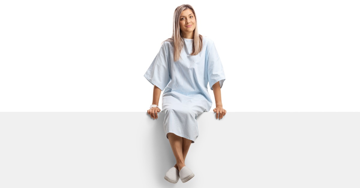 A woman wearing a light blue hospital gown and white slippers sitting on a white panel with her ankles crossed.