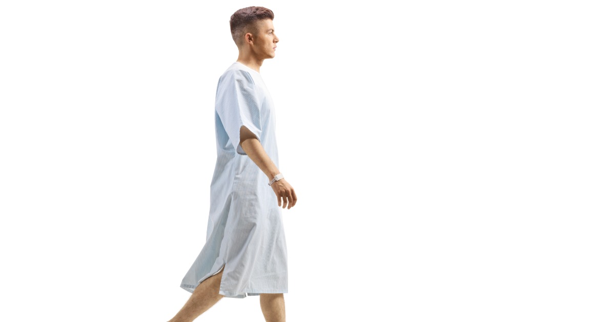 A woman wearing a light blue hospital gown and white slippers sitting on a white panel with her ankles crossed.